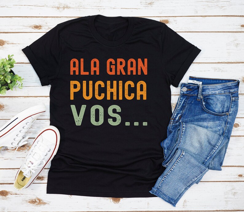 May include: Black t-shirt with a vintage-style graphic that reads "ALA GRAN PUCHICA VOS..." in orange, yellow, and green text.