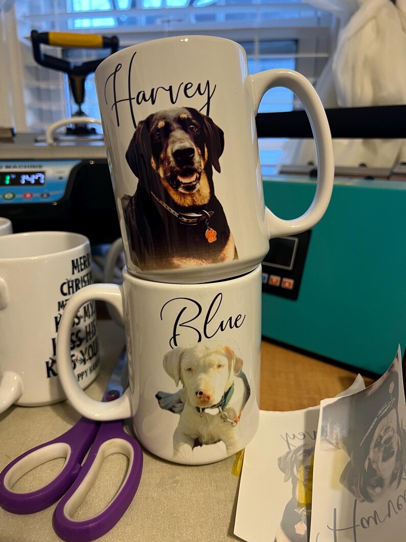 May include: Two white ceramic mugs with dog photos and names. The top mug has a black dog named Harvey and the bottom mug has a white dog named Blue.