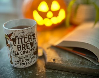 May include: A white ceramic mug with a black and white design that says "Witch's Brew Tea Company" and "Have a cup and sit for a spell". The mug is filled with dark tea and is sitting on a wooden surface in front of a lit jack-o'-lantern.