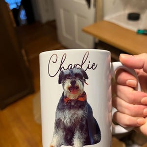 May include: White ceramic mug featuring a black and grey dog wearing a red and black bow tie. The dog's name, "Charlie", is written in cursive on the mug.