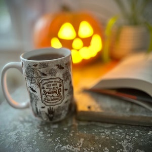 Puede incluir: Una taza de cerámica blanca con un diseño en blanco y negro que presenta murciélagos, telarañas y el texto "Sanderson Sisters Est 1693 Brewing Co. Salem Mass." La taza está llena de un líquido oscuro y está sentada en una mesa junto a un libro y una calabaza iluminada.