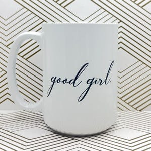 Può includere: Una tazza in ceramica bianca con una scritta in corsivo nero che dice "good girl".
