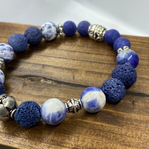 May include: A bracelet with a mix of blue and white stone beads, dark blue lava beads, and silver-coloured metal accents. The bracelet is strung on a stretchy cord. The bracelet is displayed on a wooden surface.