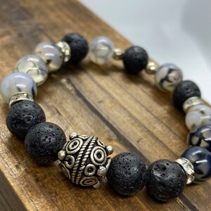 Peut inclure: Un bracelet de perles composé de perles de lave noire, de perles grises et blanches marbrées et de perles d'accent argentées. Le bracelet comprend une perle décorative argentée avec un motif complexe. Le bracelet est présenté sur une surface en bois.