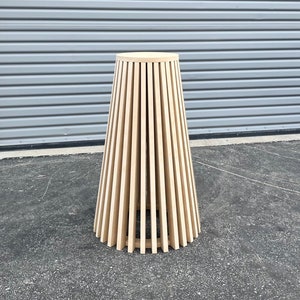 May include: A wooden table with a round top and a unique design. The table is made of thin, vertical slats of wood that create a cone shape. The table is standing on a gray asphalt surface.