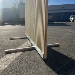 May include: A wooden room divider with vertical slats, supported by a silver metal base. The natural wood tones contrast with the metallic base, creating a modern aesthetic. The divider is designed for privacy or to section off a space.