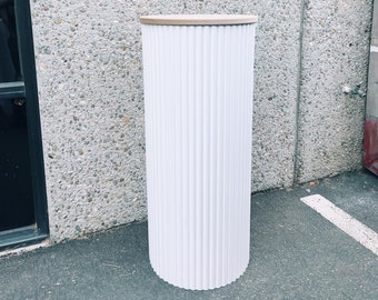 May include: A tall, white, fluted pedestal with a light brown top. The cylindrical column has vertical grooves and a scalloped base. The pedestal is set against a textured gray wall and a concrete surface.