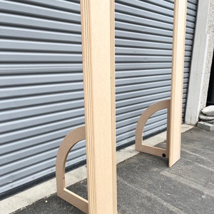 May include: Two unfinished wooden archways with rounded tops, standing upright on a gray asphalt surface. The archways are made of plywood and have a simple, modern design.