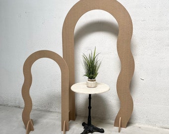 May include: Two cardboard arches, one larger than the other, with a wavy design. The arches are standing on a concrete floor. A small round table with a white top and black legs is in front of the arches. A small green plant is on the table.