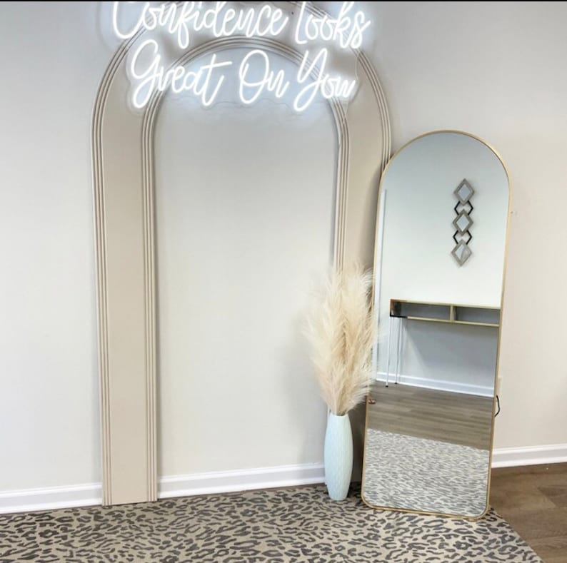 May include: A white neon sign with the text "Confidence looks great on you" is mounted on a white wall with a large arched doorway. A tall, gold-framed mirror with an arched top is in the foreground. A vase with dried pampas grass is in front of the mirror.