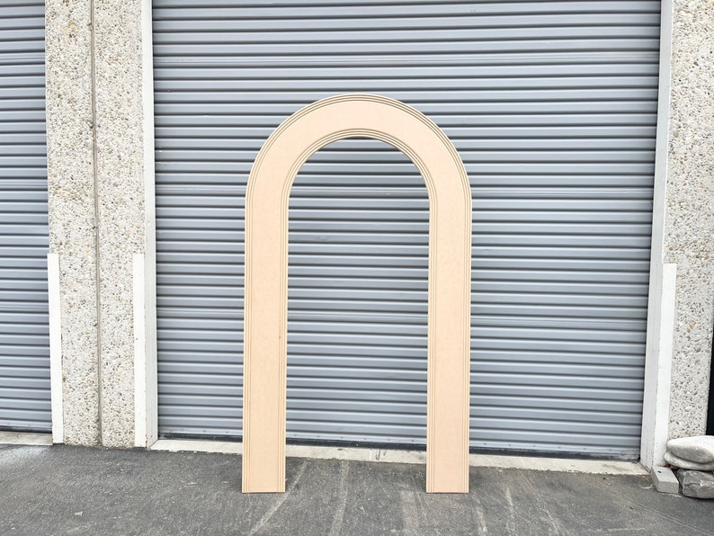 May include: A wooden archway, made of plywood, with a rounded top and a natural wood finish. The archway is standing upright against a gray metal garage door.