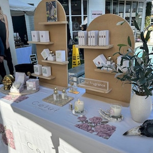 May include: A white table with a white tablecloth featuring a floral design. On the table are two wooden display stands with shelves, showcasing various products, including candles, bottles, and boxes. The stands are decorated with the text "Celui Fragrance".