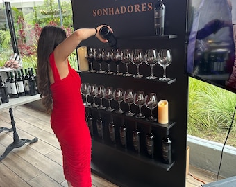 May include: A black arched display stand features the text THE SONHADORES with a tree logo. It holds rows of wine glasses and bottles on shelves. A person in a red dress pours wine into a glass. Two pillar candles are placed on the shelves.