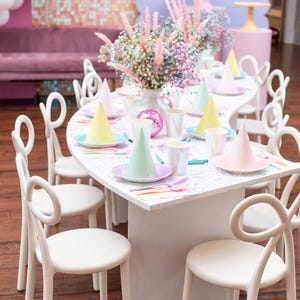 May include: A children's party table set with pastel party hats, plates, and cups. The table is decorated with a floral centrepiece and surrounded by white chairs. The background includes a pink sofa and a decorative wall.