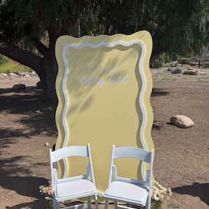 Könnte beinhalten: Ein großer, hellgelber, rechteckiger Hintergrund mit gewellten Kanten und dem weißen Schriftzug "WELCOME Baby Girl" in der Mitte. Zwei weiße Klappstühle mit gepolsterten Sitzen stehen davor, flankiert von kleinen Blumenarrangements.