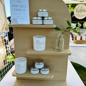 May include: A wooden display stand with three shelves, showcasing a variety of candles and wax melts. The candles are in white jars with silver lids and feature the text "Vidas" and "Memories of Heaven". The wax melts are in small silver containers with the text "Vidas" and "Kiss of Sunshine". The stand is decorated with a small vase of greenery.