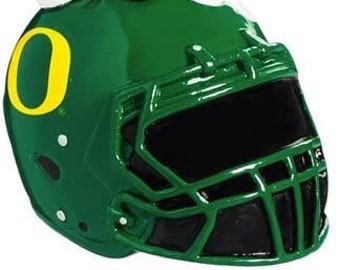 May include: Green and yellow Christmas ornament featuring an elf painting a green football helmet with the letter "O" on the side. The elf is holding a paintbrush and a can of paint that says "Pain".