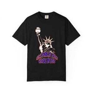 Op de afbeelding: Een zwart t-shirt met korte mouwen en een afbeelding van het Vrijheidsbeeld in witte en oranje tinten. Onder het beeld staat op de borst de tekst "NEW YORK IS ALIVE" en "KNICKS IN FIVE" in blauwe letters met een oranje omlijning.
