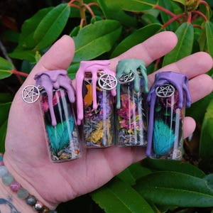 Peut inclure: Quatre petits flacons en verre avec des contenus colorés et des dessus en cire dégoulinante violet, rose et vert. Chaque flacon a un charme argenté avec un pentagramme. Les flacons sont tenus dans une main sur un fond de feuillage vert.