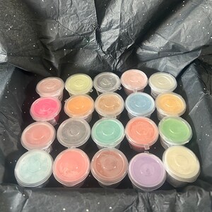 May include: A collection of 20 small, round containers filled with colourful wax. The containers are arranged in a grid pattern and are made of clear plastic. The wax is a variety of colours, including pink, blue, green, orange, and yellow.
