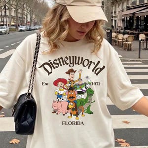 Può includere: T-shirt color crema con grafica Disneyworld con personaggi dei cartoni animati e la scritta "Disneyworld Est 1971 Florida". La maglietta è abbinata a un berretto da baseball beige e una borsa a tracolla trapuntata nera.