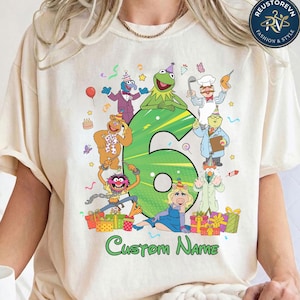 Könnte beinhalten: Ein cremefarbenes T-Shirt mit einer großen grünen Zahl sechs, umgeben von verschiedenen Muppet-Figuren mit Partyhüten, darunter Kermit, Miss Piggy und Fozzie, mit bunten Geschenken am unteren Rand und dem Text Custom Name.