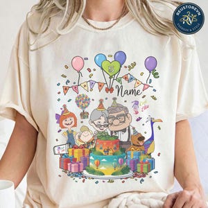 May include: A cream-colored t-shirt featuring a colorful graphic of characters from the movie Up, including an elderly man, a young boy, and a bird. The design includes a two-tiered birthday cake, balloons, gift boxes, and the text "5 Name".