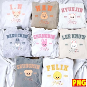 Puede incluir: Una colección de ocho sudaderas con capucha en varios colores, cada una con un nombre en fuente universitaria, un año de nacimiento, un personaje animal de dibujos animados y el logotipo de Stray Kids. Las sudaderas son blancas, beige, grises y crema.