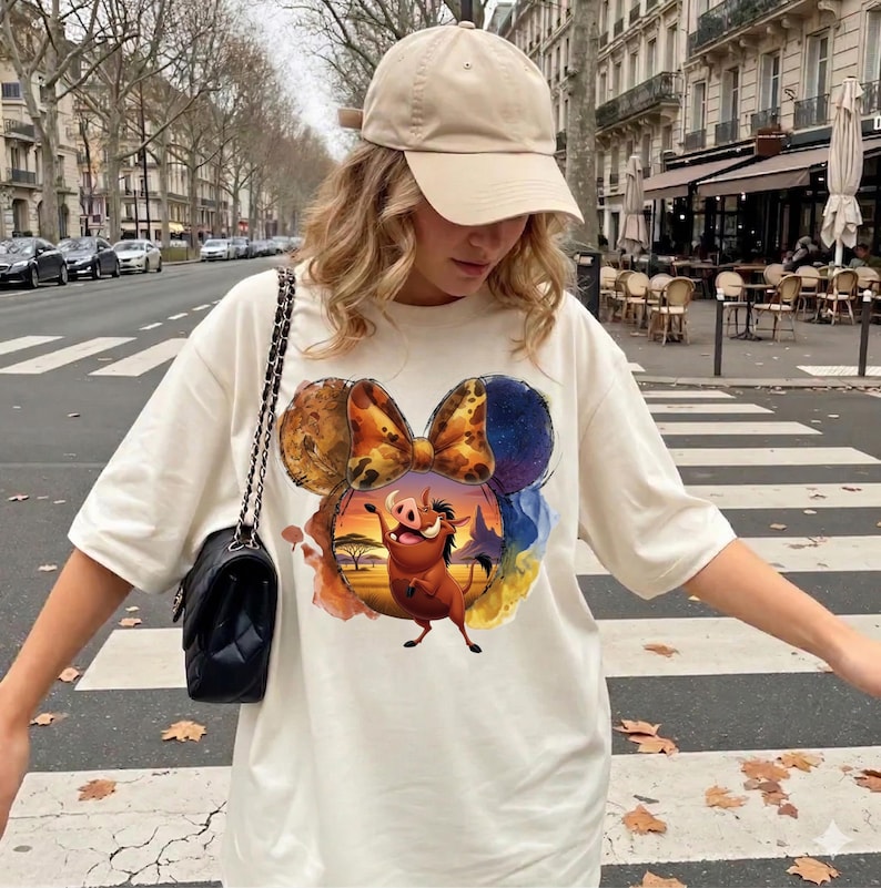 May include: Cream-colored t-shirt featuring a colorful graphic of a cartoon character in a sunset scene, framed by Mickey Mouse ears. The shirt is paired with a beige baseball cap and a black shoulder bag. The background shows a city street.