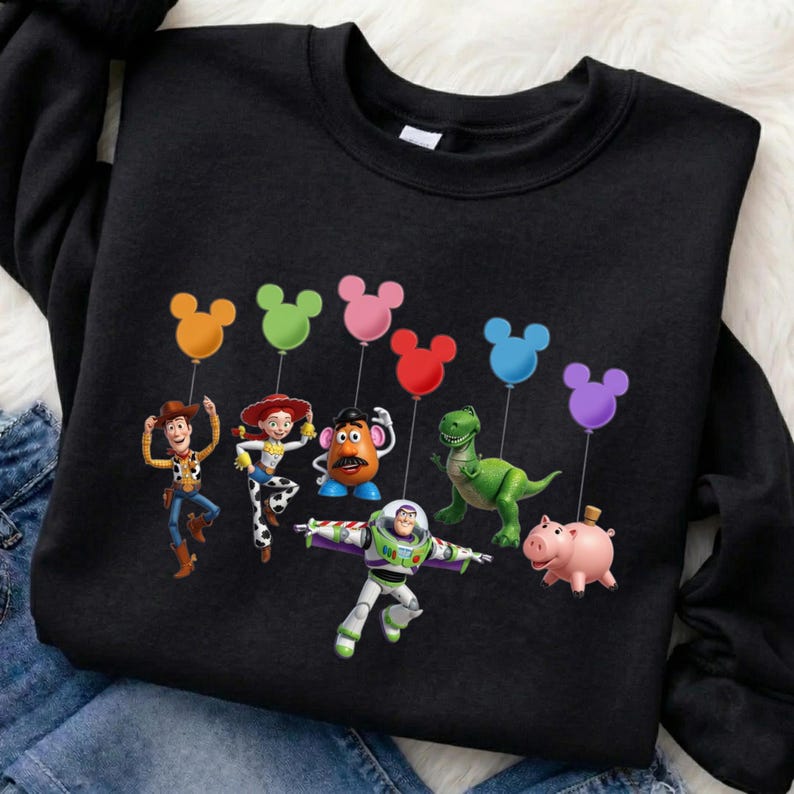 Puede incluir: Sudadera negra con personajes de Toy Story, cada uno sosteniendo un globo con forma de Mickey Mouse. Los personajes incluyen a Woody, Jessie, Sr. Cara de Papa, Rex, Buzz Lightyear y Hamm.