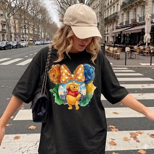 May include: Black t-shirt featuring a colorful graphic of Winnie the Pooh with a bow and the word "Pooh". The design includes a cityscape and various colors. The shirt is a loose fit.
