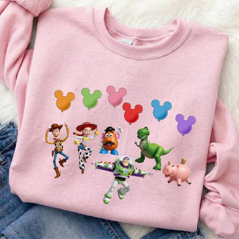 Puede incluir: Una sudadera rosa claro con un gr&aacute;fico de personajes de Toy Story, cada uno sosteniendo un globo con forma de Mickey Mouse. Los personajes incluyen a Woody, Jessie, Sr. Cara de Papa, Buzz Lightyear, Rex y Hamm. La sudadera es de material suave.