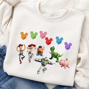 Puede incluir: Sudadera blanca de cuello redondo con un gr&aacute;fico colorido de personajes de Toy Story. Los personajes sostienen globos con forma de Mickey Mouse de varios colores. El dise&ntilde;o incluye a Woody, Jessie, Sr. Potato, Buzz Lightyear, Rex y Hamm.