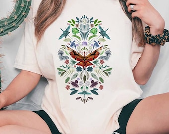 Pode incluir: Camiseta creme com um design simétrico de uma mariposa vermelha e laranja com as asas abertas, rodeada por elementos botânicos verdes e azuis. O design inclui pássaros estilizados e padrões florais.