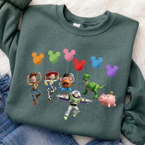 Puede incluir: Una sudadera verde oscuro con un gr&aacute;fico colorido de personajes de Toy Story. Los personajes sostienen globos con forma de Mickey Mouse en naranja, verde, rosa, rojo, azul y morado. La sudadera es de material suave.