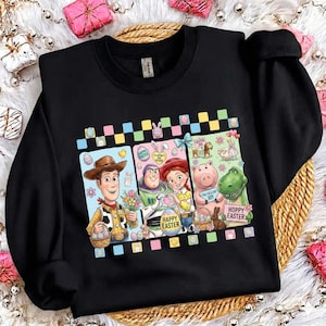 Puede incluir: Sudadera negra con un gráfico colorido de los personajes de Toy Story celebrando la Pascua. Woody, Buzz Lightyear, Jessie, Hamm y Rex se representan con huevos de Pascua y las palabras "Happy Easter" y "Hoppy Easter".
