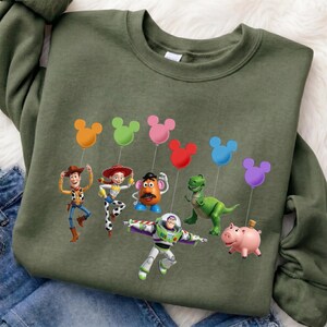 Puede incluir: Una sudadera verde oliva con un gr&aacute;fico colorido de los personajes de Toy Story. El dise&ntilde;o incluye a Woody, Jessie, Sr. Potato, Rex, Buzz Lightyear y Hamm, cada uno sosteniendo un globo con forma de Mickey Mouse en varios colores.