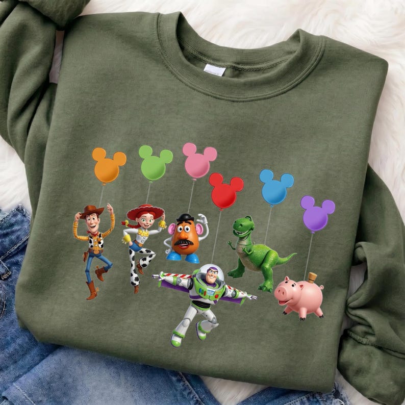 Puede incluir: Una sudadera verde oliva con un gr&aacute;fico colorido de los personajes de Toy Story. El dise&ntilde;o incluye a Woody, Jessie, Sr. Potato, Rex, Buzz Lightyear y Hamm, cada uno sosteniendo un globo con forma de Mickey Mouse en varios colores.