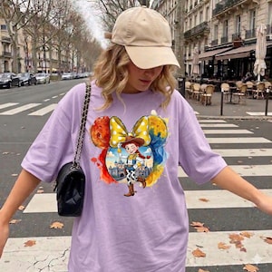 May include: A light purple t-shirt featuring a colorful graphic of a cartoon character in a cowboy hat and a yellow polka dot bow. The shirt is paired with a beige baseball cap and a black shoulder bag.