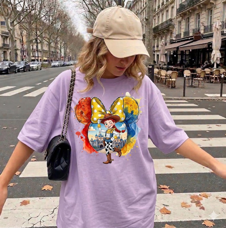 May include: A light purple t-shirt featuring a colorful graphic of a cartoon character in a cowboy hat and a yellow polka dot bow. The shirt is paired with a beige baseball cap and a black shoulder bag.