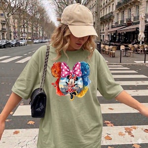 May include: Sage green t-shirt featuring a colorful Minnie Mouse graphic. The design includes a large pink bow with white polka dots, and a cartoon image of Minnie Mouse in a red dress. The shirt is paired with a tan baseball cap and a black shoulder bag.