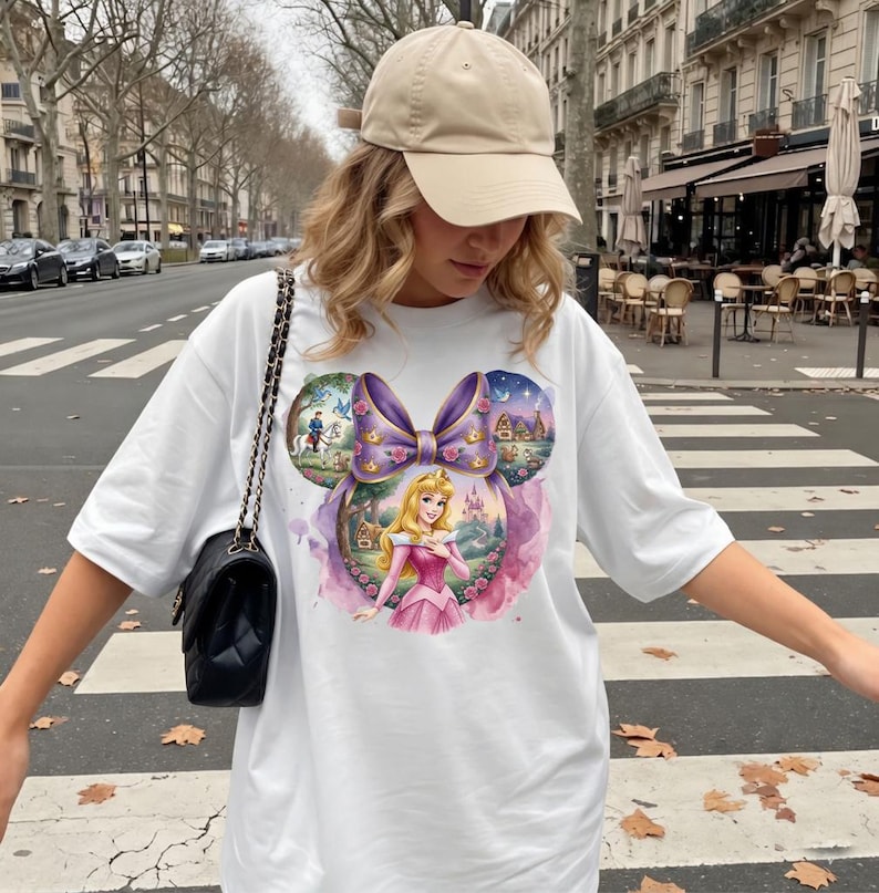 May include: White t-shirt featuring a colorful graphic of Princess Aurora in a pink dress, set within a purple bow-shaped design. The design includes elements of a castle, forest, and a prince on a horse. The shirt is worn with a beige baseball cap.