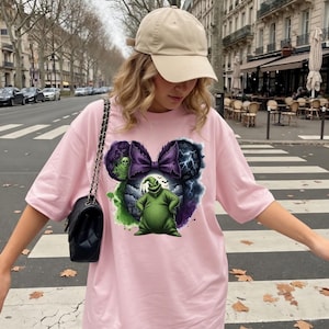 May include: A light pink t-shirt featuring a graphic of a green character with a purple bow and lightning bolts. The design is in the shape of Mickey Mouse ears. The shirt is worn with a tan baseball cap and a black purse.