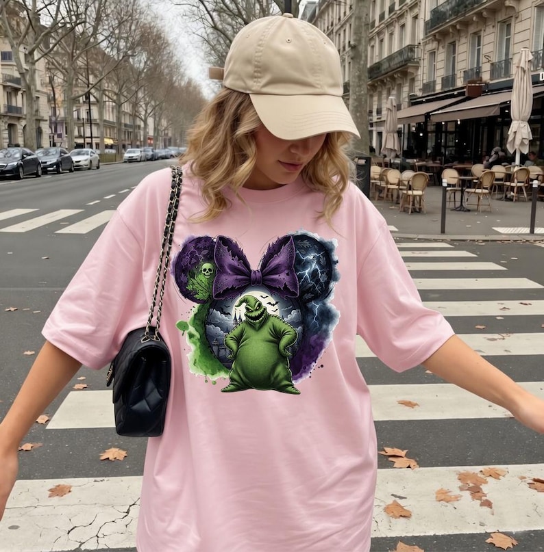 May include: A light pink t-shirt featuring a graphic of a green character with a purple bow and lightning bolts. The design is in the shape of Mickey Mouse ears. The shirt is worn with a tan baseball cap and a black purse.