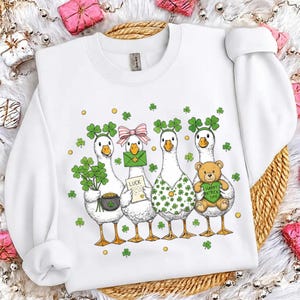 May include: White sweatshirt with a St. Patrick's Day theme, featuring four cartoon geese. Each goose is adorned with clovers, a pot of gold, a letter with the word "LUCK", or a teddy bear holding a heart that says "Happy Patrick Day".