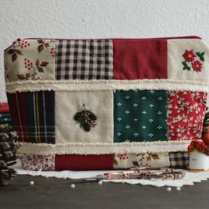 May include: A patchwork fabric zippered pouch with a red, green, and white color scheme. The pouch is decorated with embroidered pine cones and holly berries.