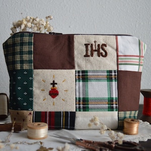 May include: A brown and green patchwork fabric zippered pouch with embroidered details. The pouch has a cross and a heart on one side and the letters "IHS" on the other side.