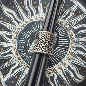 May include: A wide silver-toned ring featuring an intricate, repeating openwork floral or geometric lattice pattern. The band has a slightly concave, hourglass-like shape, narrowing in the center and widening toward the edges, with a polished finish.
