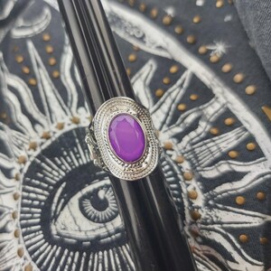 May include: A silver-toned ring featuring an oval-shaped, smooth purple cabochon stone set within an intricate, textured metal frame. The setting displays a decorative border with fine bead-like detailing surrounding the central purple stone.