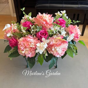 May include: A floral arrangement with large pink peonies, smaller pink roses, and white flowers with green leaves. The arrangement is in a green metal container. The text "Marlene's Garden" is at the bottom.